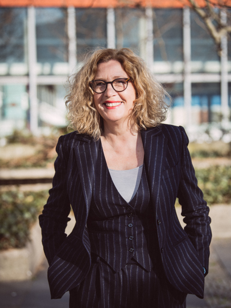 Martina Löw stands in a suit and glasses facing the camera with her hands in her pockets.
