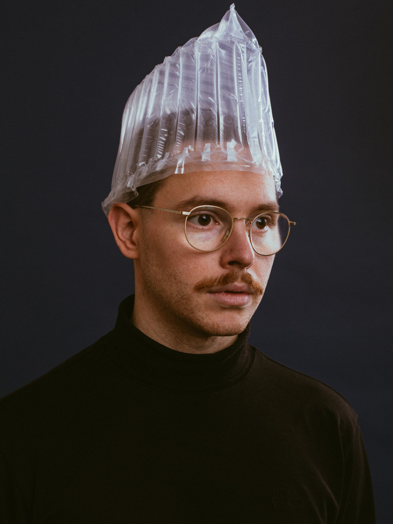 Porträt des Berliner Illustrators Felix Bork, er trägt ein schwarzes Oberteil, eine durchsichtige Haube auf seinem Kopf und steht vor einen schwarzen Hintergrund.