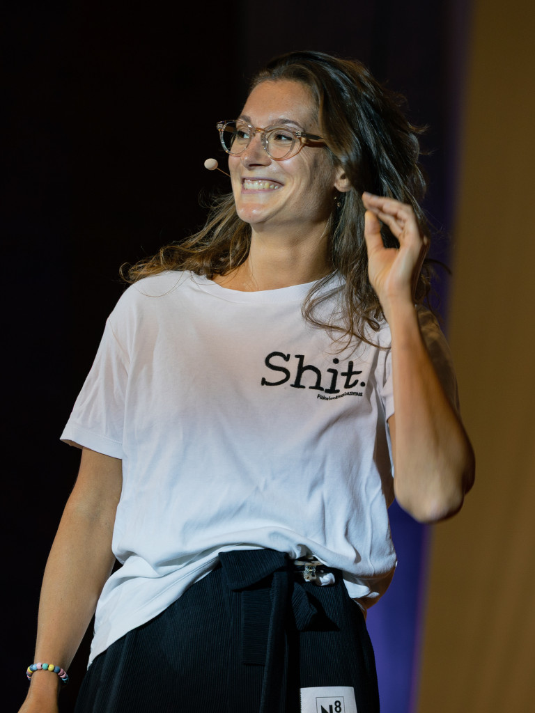 Lisa Budzinski (Biotechnologin und Moderatorin) auf einer Bühne vor großem Publikum, sie trägt helle Kleidung und zeigt mit dem Finger auf etwas außerhalb des Bildes.