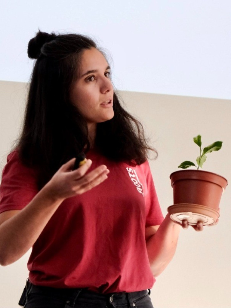 A woman holding a flowerpot with a small green plant in her left hand, explaining something