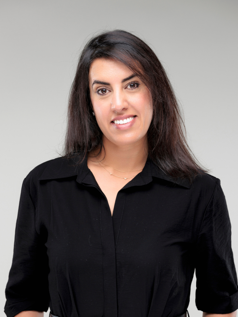 Grey background, Anjam is looking straight, long hairs, smiling, wearing a black shirt