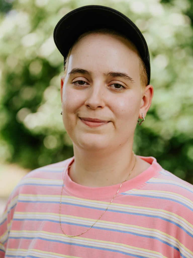 A foto of Clara Isakowitsch smiling into the camera, wearing a pink shirt and a cap. In the background are green bushes, blurred.