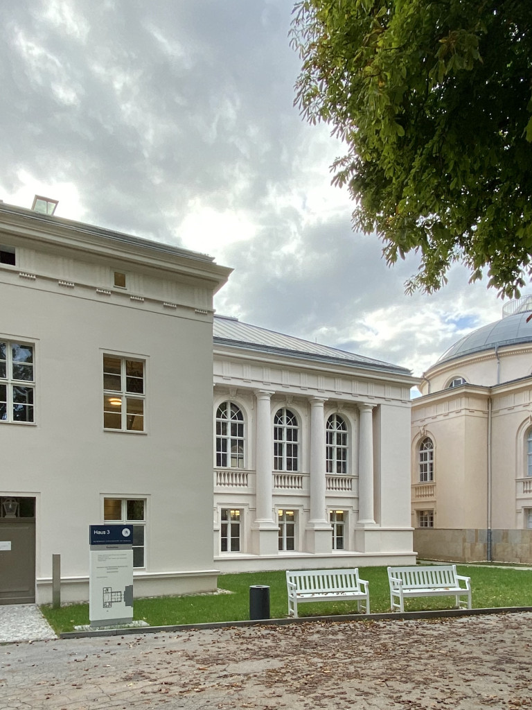 Exterior view of the Hermann von Helmholtz-Zentrum für Kulturtechnik at Humboldt-Universität zu Berlin, with the Tieranatomisches Theater’s domed architecture on the right. A courtyard with benches, a bicycle, and a large tree are in the foreground.