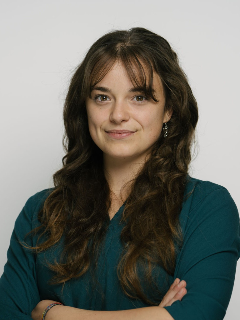 A portrait of a women in a dark green long sleeve. Her upper body is seen standing in front of a white wall, her arms are crossed. She has long brown hair and smiles softly.