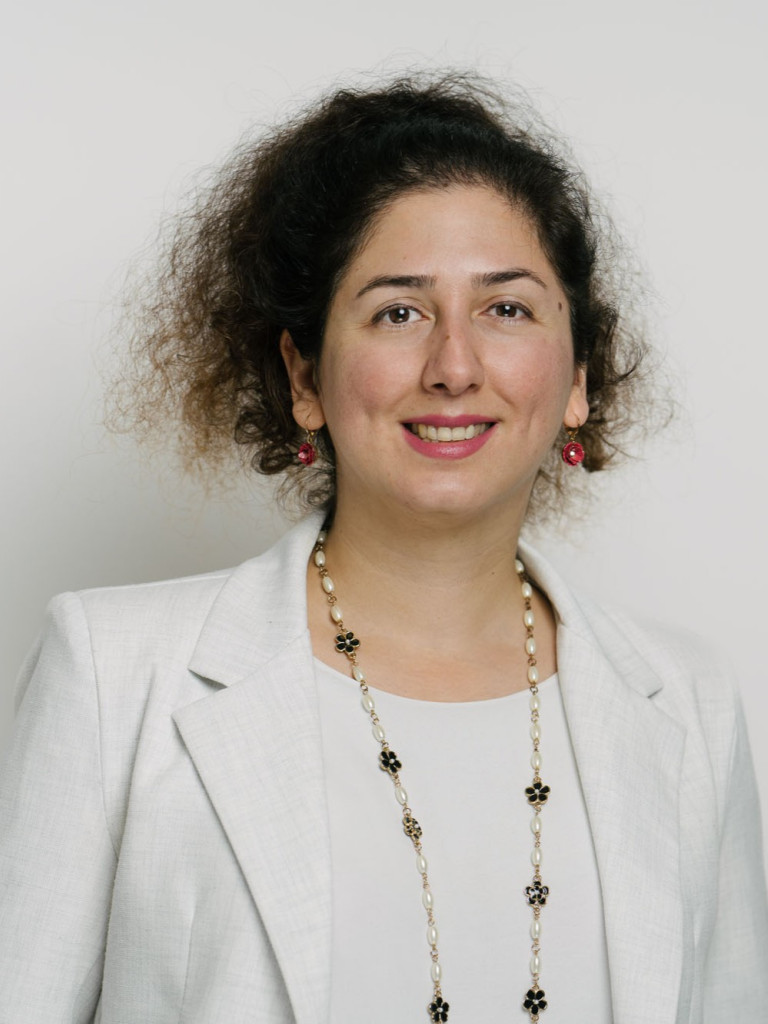 A portrait of the upper half of a women standing in front of a white background. She is wearing a white suit and a long necklace and earings. She has curly brown hair that is done up and is smiling with her teeth.
