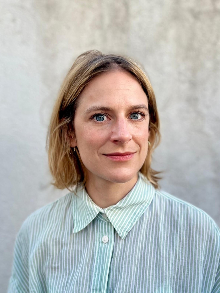 A women with short light brown to blonde hair in front of a grey wall. The portrait shows her face and body just until the mid of her upper arms. She is wearing a button down striped shirt in blue with and is slighty smiling without her teeth showing.