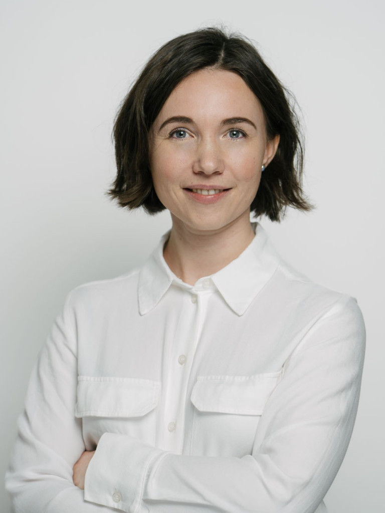 A portrait of a women that stands with her arms crossed in front of a white wall. She has short brown hair and is smiling.