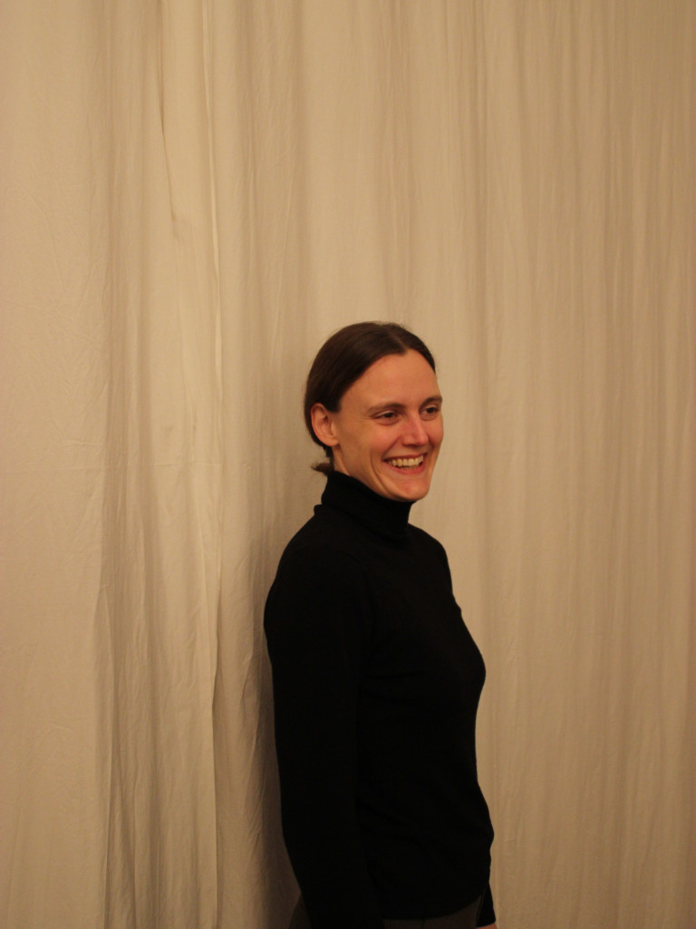 Frau Steht lächelnd vor einem weißen Vorhang. Sie trägt einen schwarzen Rollkragen Pullover.