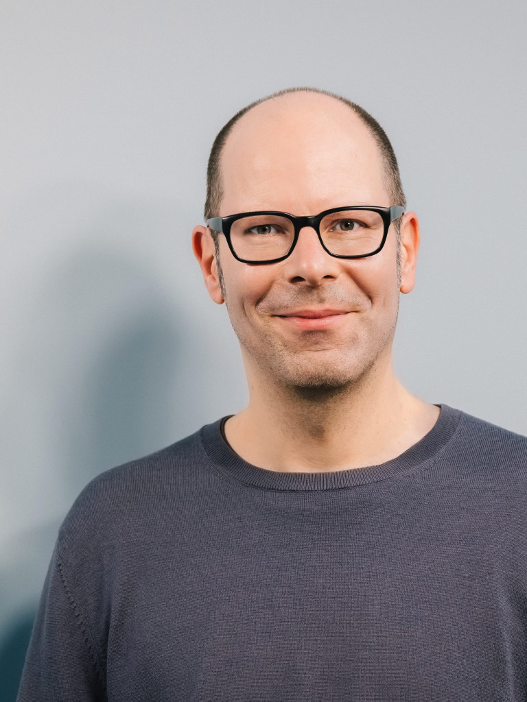 Shows a white man in grey T-Shirt wearing black glasses. 