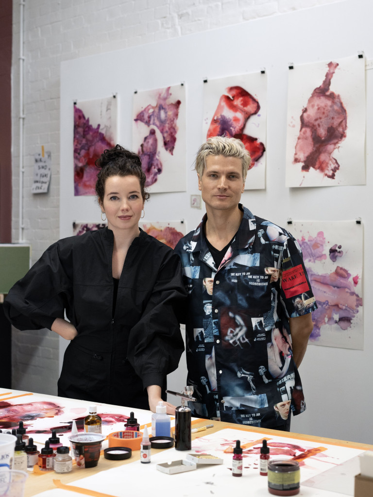 The image shows a white woman with brown hair on the left wearing a black blouse and on the right a white man with blonde hair and blue-patterned shirt. They stand in front of their watercolor artworks. 