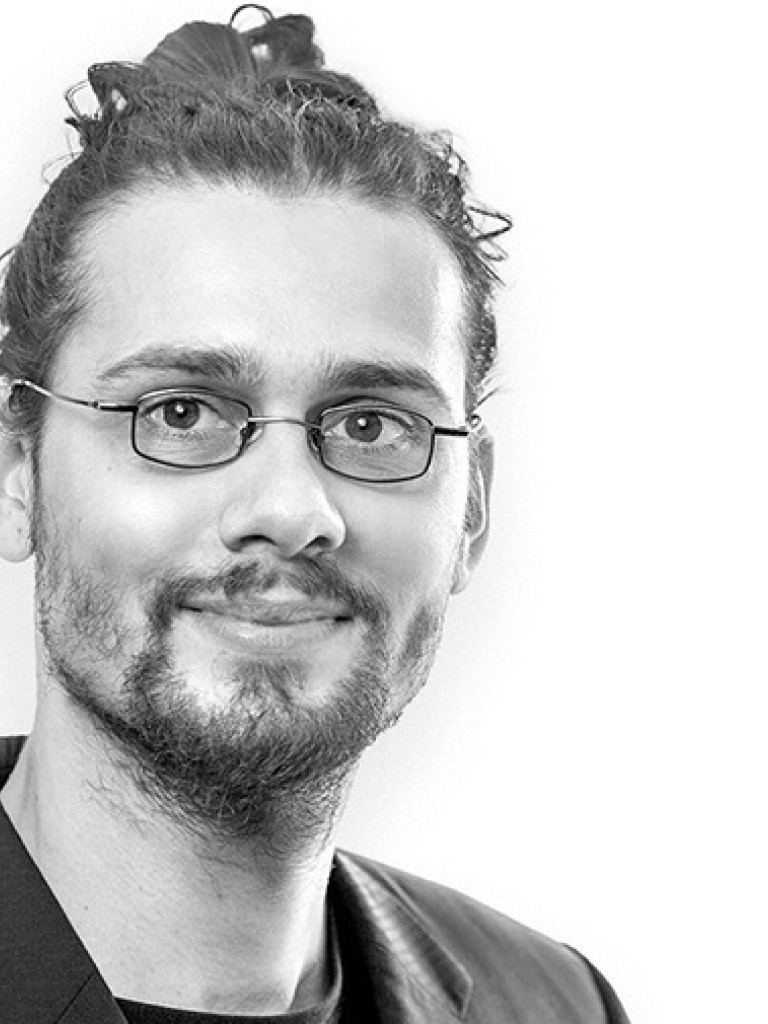 A black-and-white portrait of a person with medium-length curly hair tied in a bun. They have a short beard and mustache, and they are wearing rectangular glasses. The person is smiling slightly and dressed in a dark jacket over a light shirt. The background is plain white.