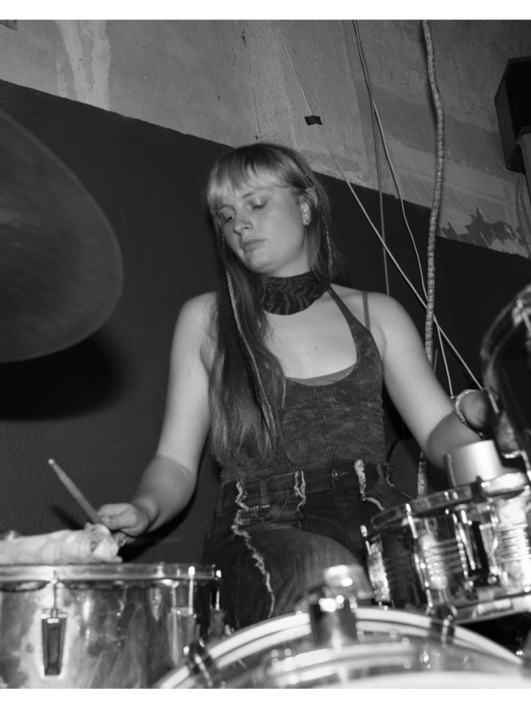 Schwarz-Weiß-Foto einer Person am Schlagzeug, die konzentriert spielt, mit langen Haaren und ärmellosem Oberteil.