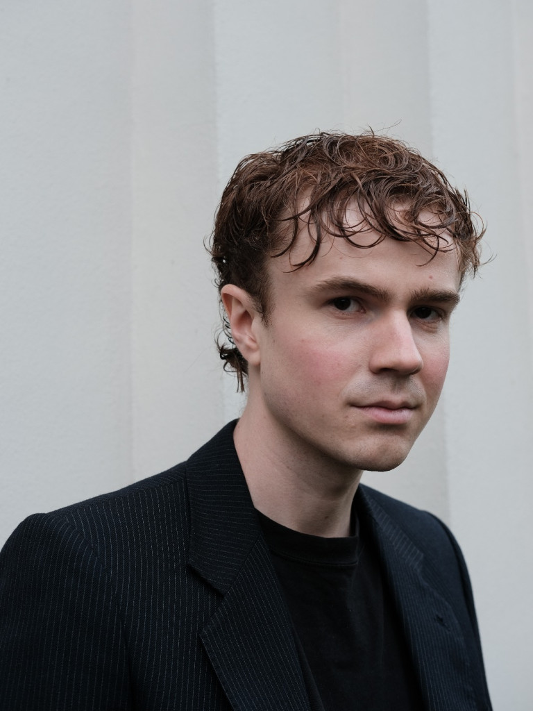 Portrait of a young person with short, wavy brown hair, wearing a black blazer and black shirt, standing against a light-colored wall.