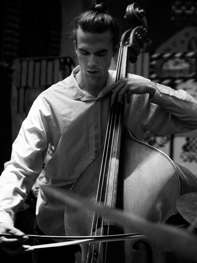 Black and white photo of a musician playing a double bass with a bow, wearing a light shirt and focusing on the instrument.