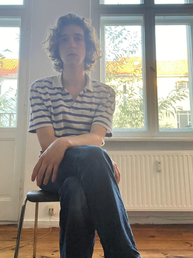 A person with curly hair wearing a striped polo shirt and jeans sits on a chair with legs crossed in front of large windows letting in daylight.