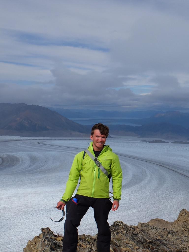 Dr. Donovan Dennis wearing a bright green jacket, standing in front of ice and snow covered landscape with mountains in the background.