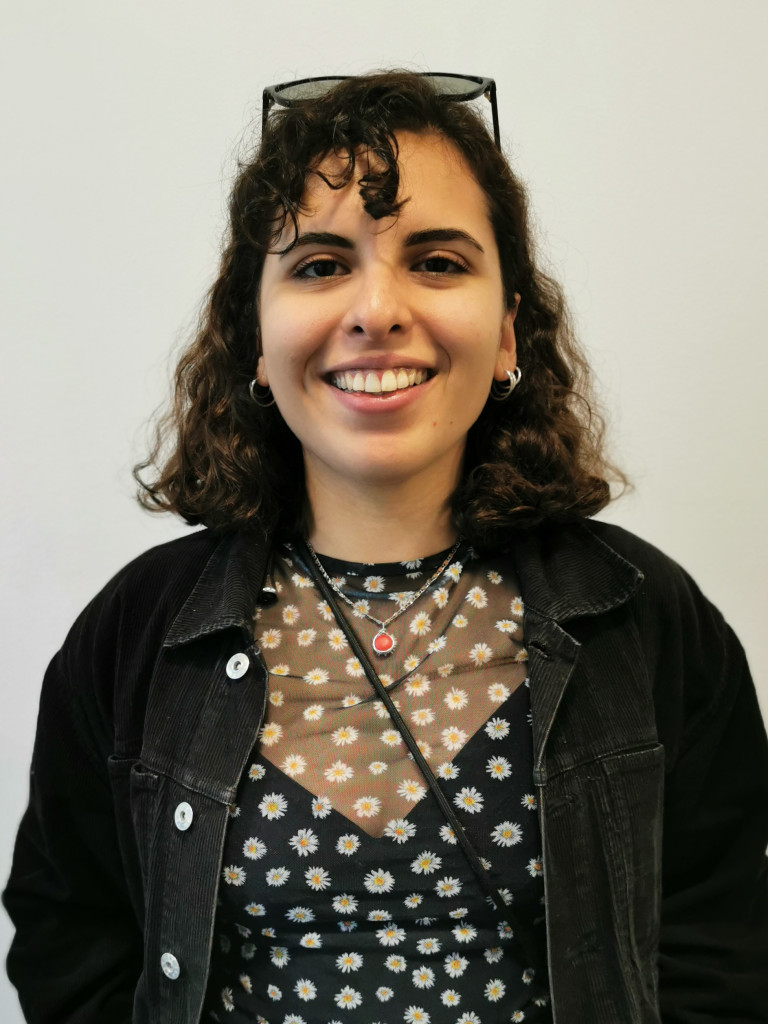 Gamze Güney smiling at the camera, wearing a back jacket and a black top. She is wearing her sunglasses on her head.