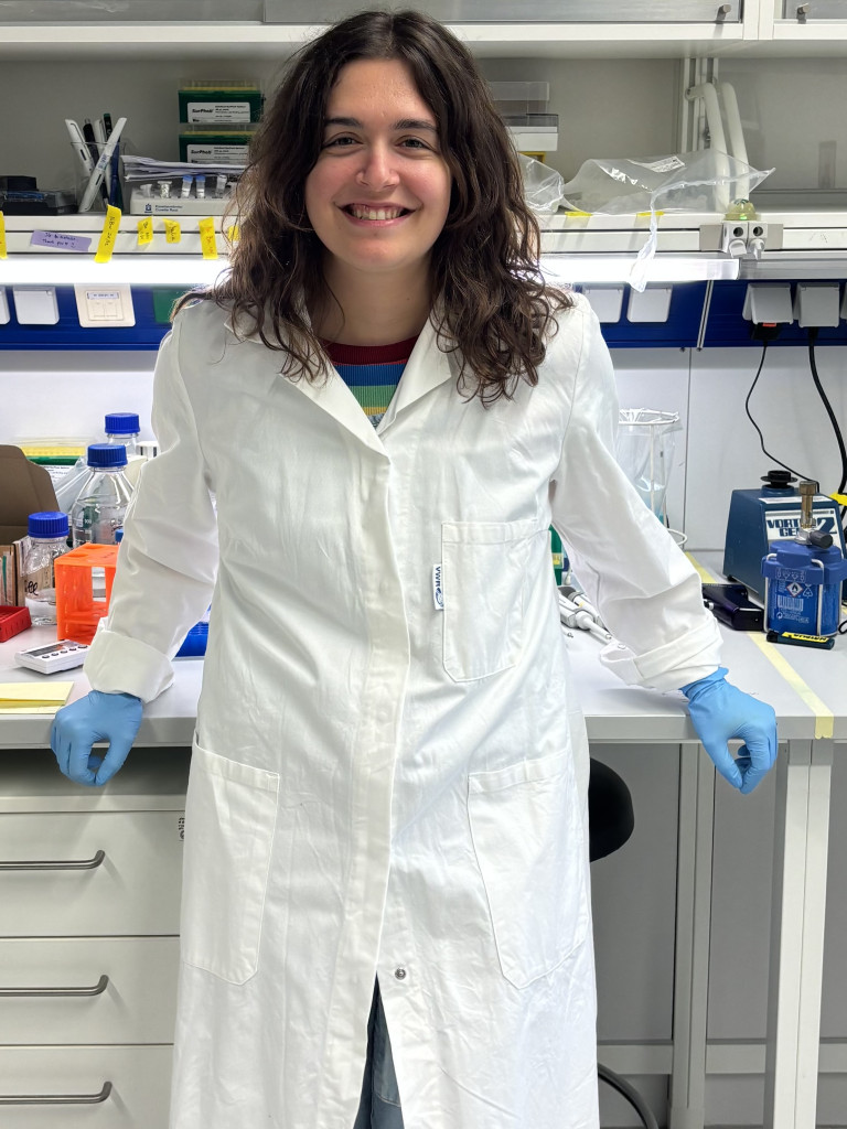 Natalia Bartzoka wearing a white lab coat and blue gloves, leaning on a desk in a laboratory.