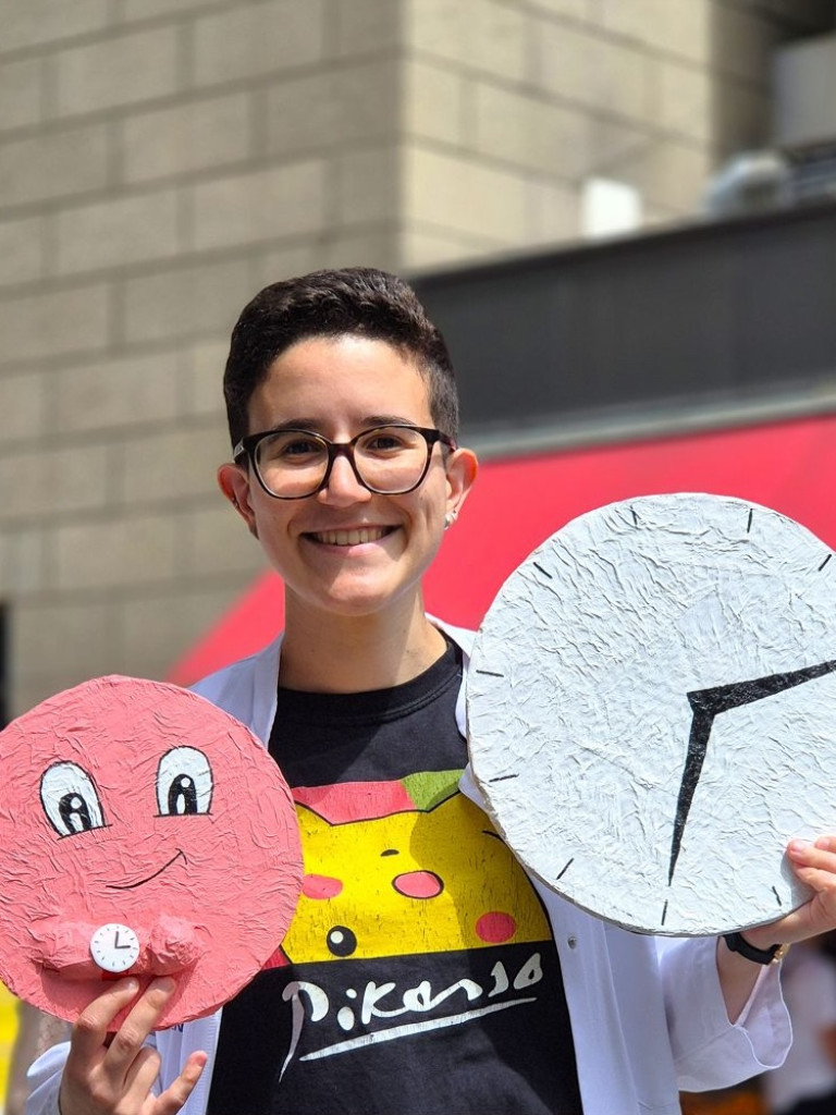 Roberta Colapietro wearing glasses, a lab coat, and a Pikachu/Picasso t-shirt, holding a prop of a clock and a smiley face with a watch.