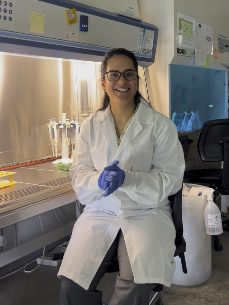 Sravanthi is wearing a white lab coat and blue gloves, sitting in her laboratory and smiling at the camera.