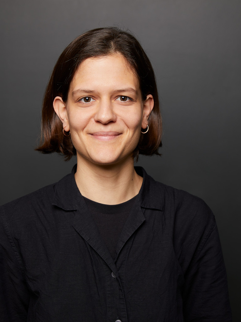 Profile picture of a women with medium-length hair, brushed back, in front of a dark background. The person is wearing a dark top and smiling slightly at the camera.