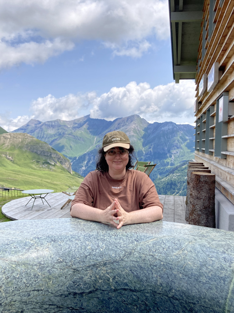 Amelia stands in the centre of the image, arms resting on a sculptural ring of rock that was once ocean crust. A scenic view of Alpine mountains is behind her. 