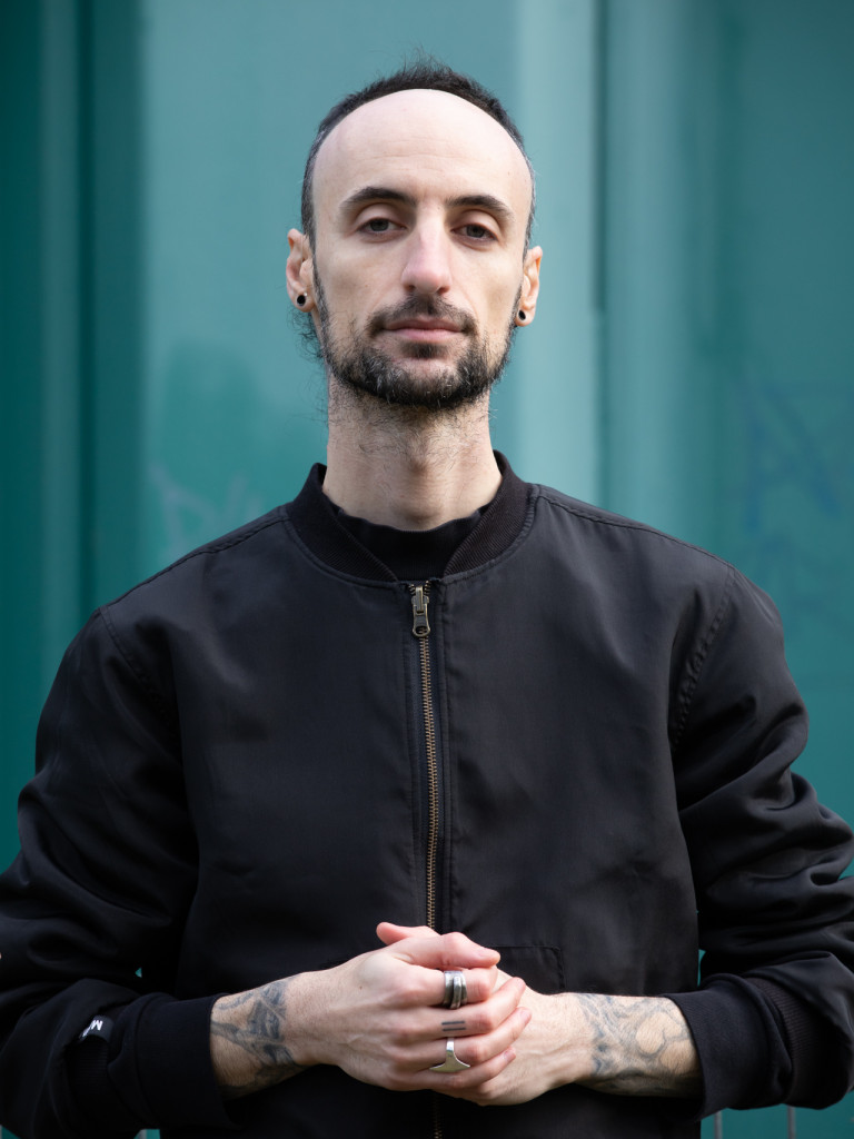 Marco Donnarumma is dressed with a black jacket and standing in front of a blue wall.