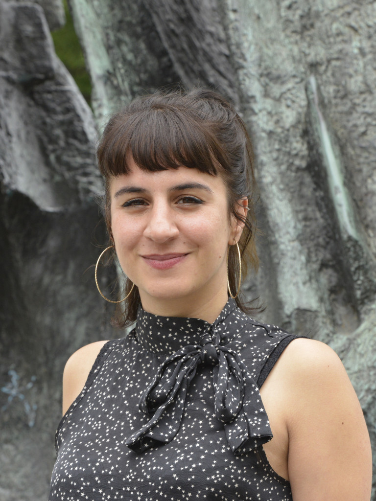 Eine weiblich gelesene Person mit dunklen Haaren steht vor einer Statue und lächelt in die Kamera.