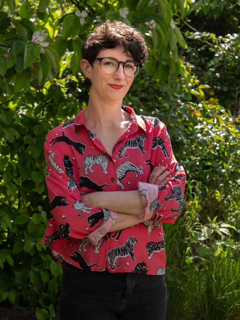 Eine weiblich gelesene Person mit kurzen Haaren steht vor einem Baum und lächelt in die Kamera. 