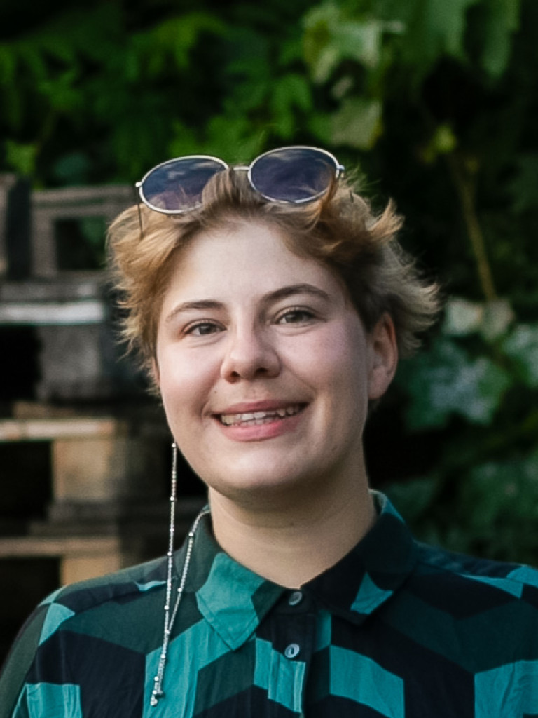 Iz is smiling into the camera. They are a white nonbinary person wearing a green checkered shirt and have their sunglasses sitting-dancing in their short blonde hair. The background is lush and full of wood and trees.