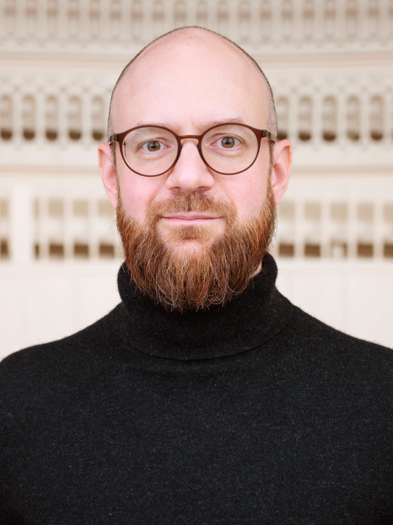 Portrait of Felix Sattler, Head Curator of the Tieranatomisches Theater, wearing round glasses and a black turtleneck, with the historic lecture theatre in the background.