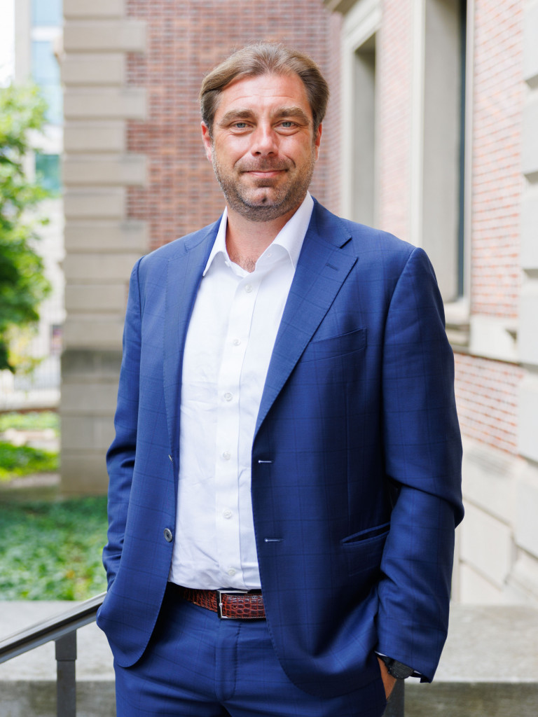 William Burke-White wearing a blue suit and open-collar white shirt stands with his hands in his pockets near the entrance of a brick building. He looks directly at the camera with a slight smile. The background shows trees, a black iron fence, and green ground cover, softly out of focus.