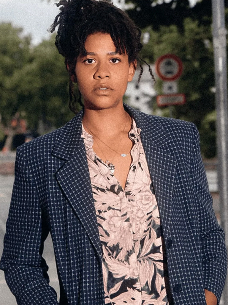 Porträt einer Person mit hochgestecktem, dunklem Lockenhaar, einer hellen, floral gemusterten Bluse und einem dunkelblau karierten Sakko vor einem unscharfen, urbanen Hintergrund mit Straßenschildern.