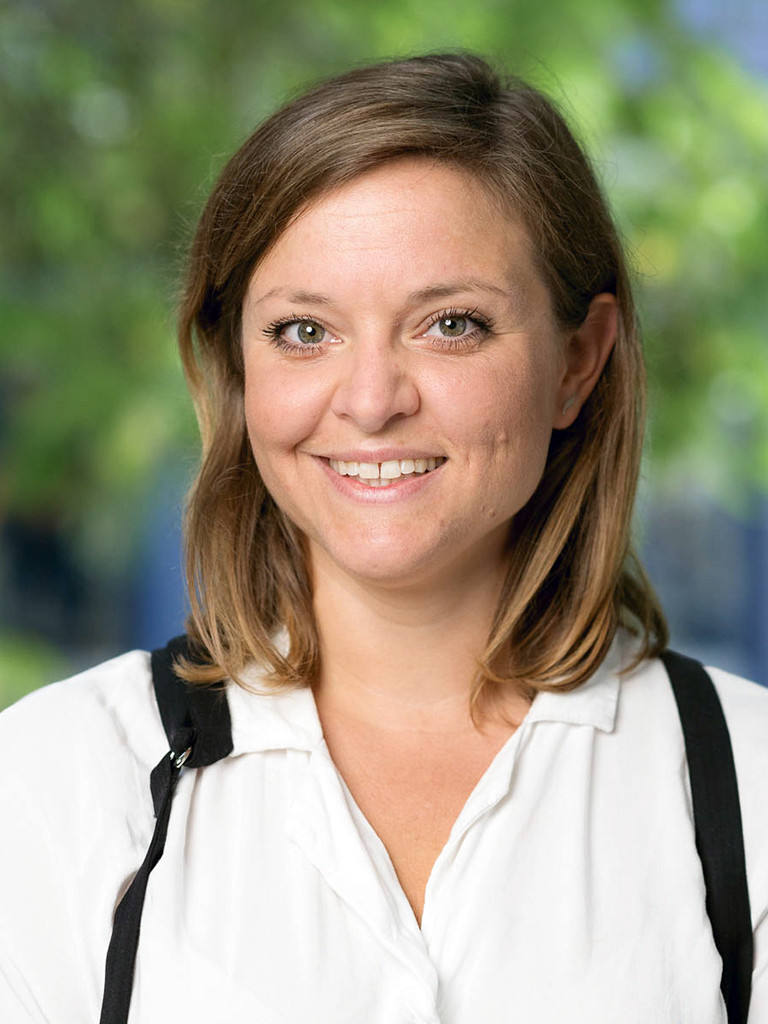 Ein Porträtfoto, das eine junge Frau mit schulterlangem Haar zeigt, die direkt in die Kamera lächelt. Sie trägt ein weißes Oberteil mit Kragen und darüber schwarze Träger. Der Hintergrund ist ein unscharfer Außenbereich mit grünem Laub.