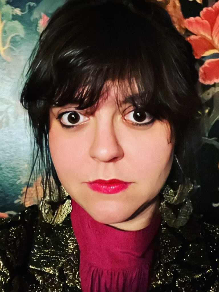 Porträt einer Person mit dunklem, gesträhntem Haar, einem leuchtend rosa Oberteil, einer schimmernden gold-schwarzen Jacke vor einem Hintergrund mit Blumenmuster.
