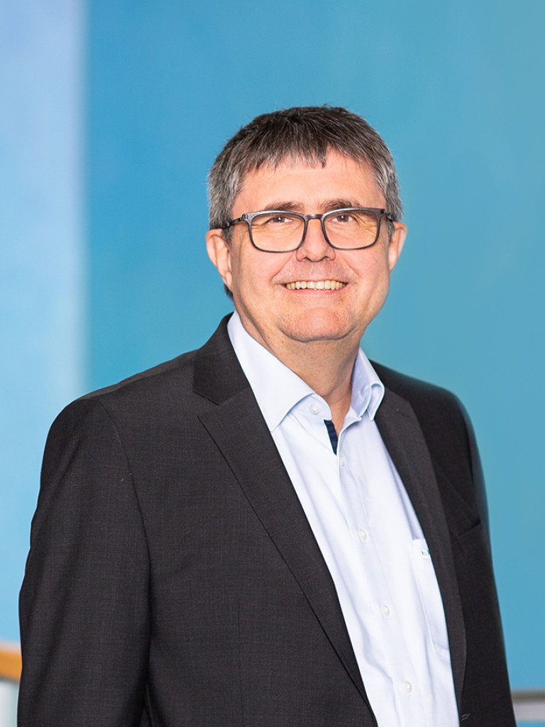 Porträtfoto von Peter Fratzl: ein lächelnder Mann mit kurzen grau-melierten Haaren und Brille, in dunklem Sakko und hellblauem Hemd, vor türkisfarbenem Hintergrund.