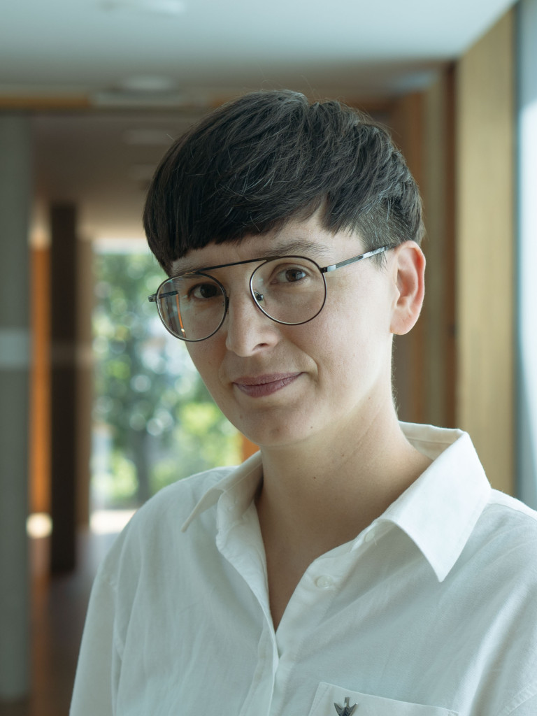 Porträt von Dr. Marianna Szczygielska: Sie hat kurze dunkle Haare und eine runde Metallbrille und blickt leicht lächelnd in die Kamera. Sie trägt ein weißes Hemd und steht in einem hellen Flur mit Holzelementen und großen Fenstern.