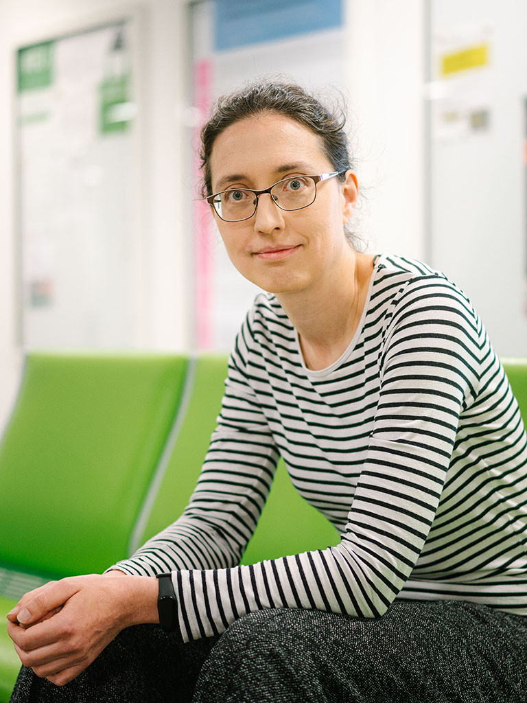 Foto einer Frau in einem gestreiften Shirt, sie sitzt auf einem grünen Sofa und trägt eine Brille.