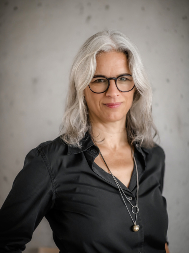 Portrait einer Frau mit grauem, schulterlangem Haar, sie trägt eine Brille.
