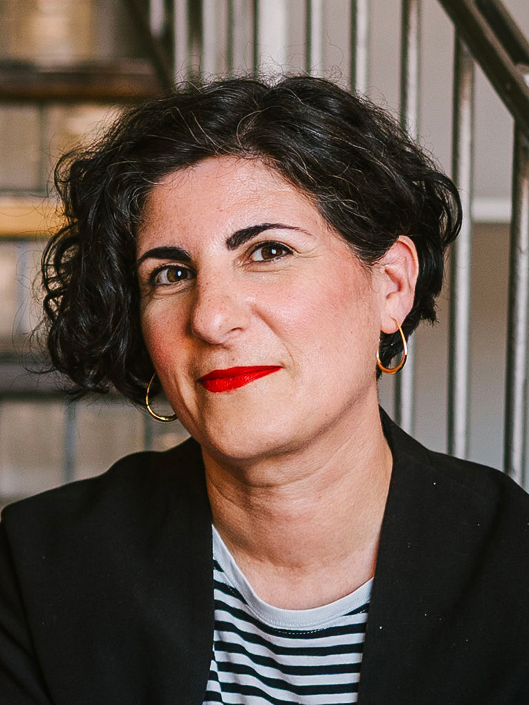 Portrait einer Frau vor einem Treppengeländer; sie hat halblanges, schwarzes Haar und trägt eine gestreiftes Shirt und einen schwarzen Blazer.