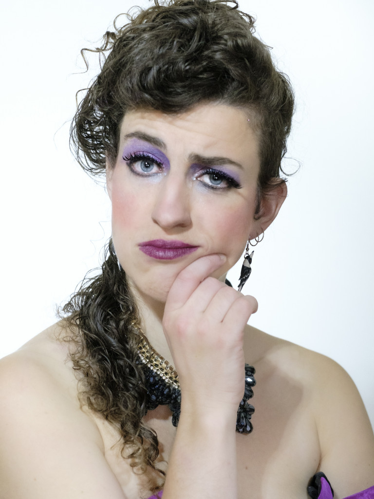 A portrait style photo of a white woman with 80s styled hair, earrings, purple makeup and dress. She puts her hand to her chin in an exaggerated thinking pose.
