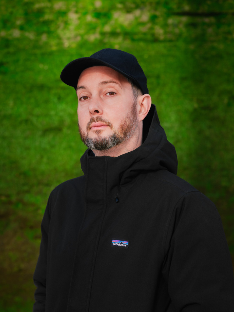 Image of artist Edwin Mingard standing against a green background. He is wearing a black baseball cap and black jacket. 