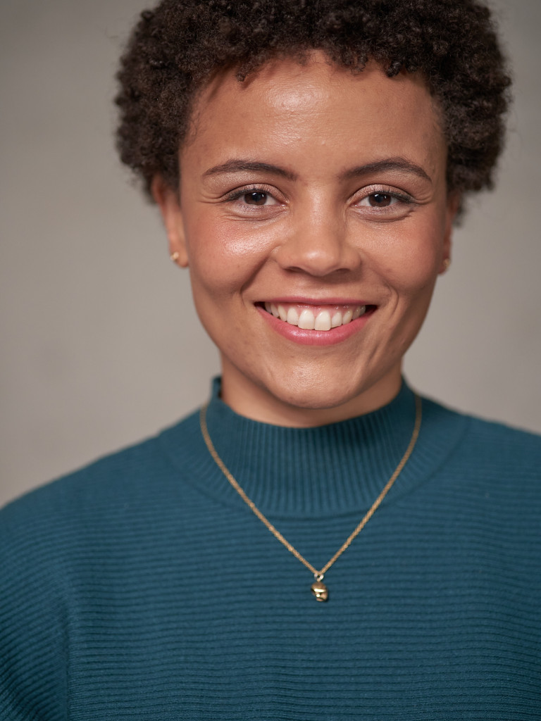 Portrait of Linda Ngono with short curly hair, wearing a teal sweater and gold necklace against a neutral background.