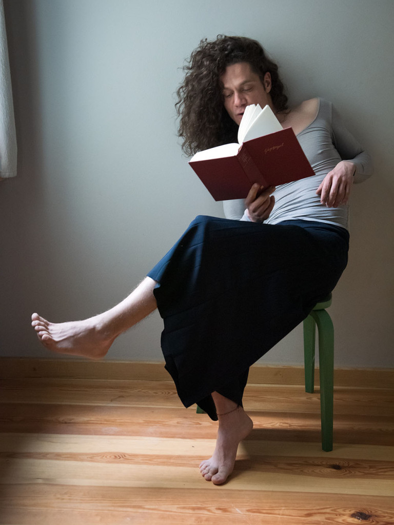 Tobias Wieland with curly hair sits barefoot on a green chair by a window, reading a red book in a softly lit room.