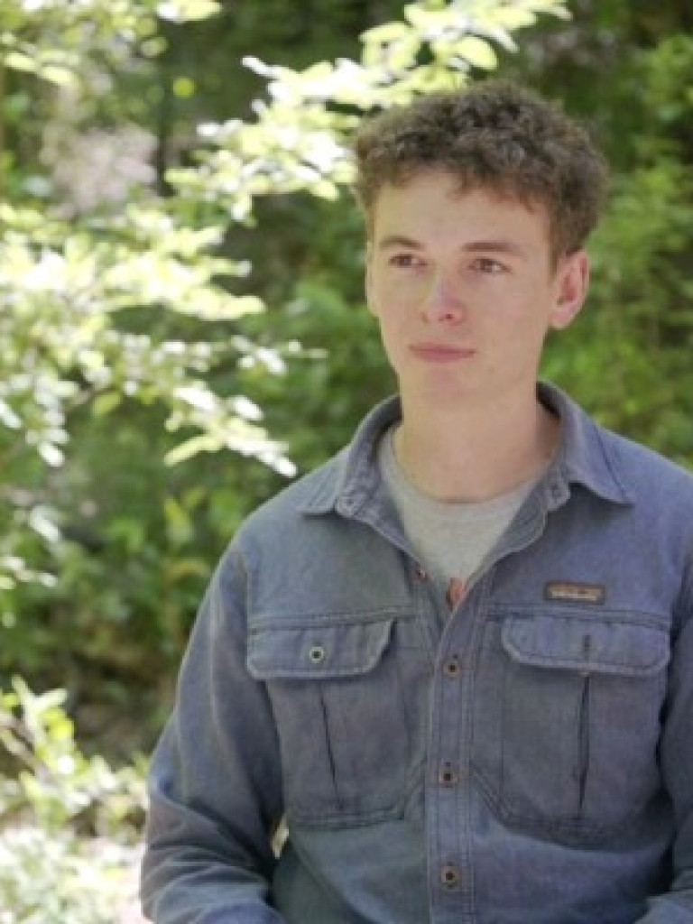 Portrait of Simon Handy wearing a blue work shirt, seated outdoors in a sunlit woodland.