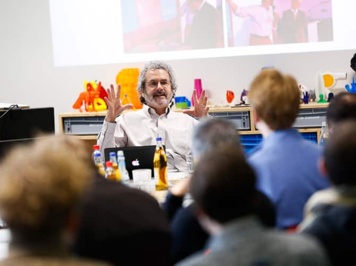 A man with curly hair and glasses enthusiastically speaks to an attentive group of people in a classroom or workshop setting. Displayed on the table in front of him are various colorful objects, a laptop, and drinks. A large screen behind him shows some images.