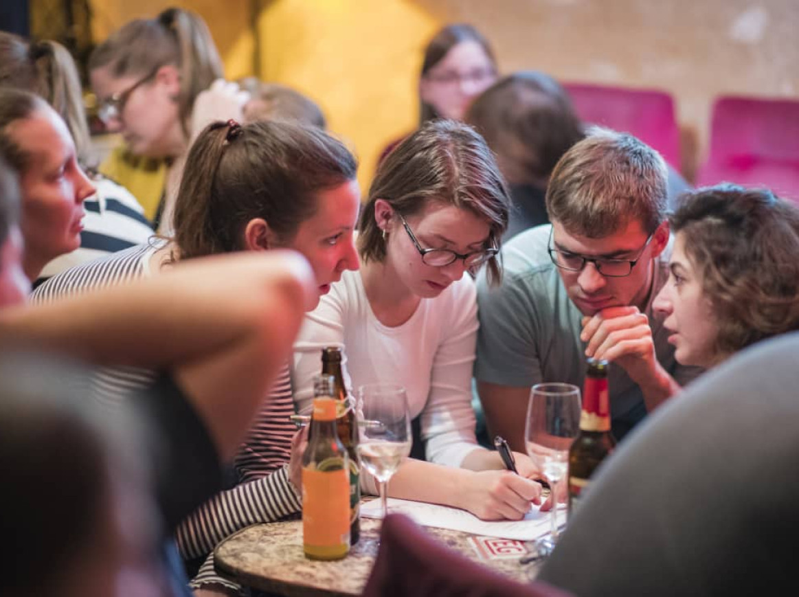 A group of people are gathered closely around a small table in a dimly lit setting, likely participating in a pub quiz or trivia night. They appear focused and engaged, with one person writing on a piece of paper while others look on, suggesting collaborative problem-solving or answering questions. Various drinks, including bottles and glasses, are present on the table, indicating a casual and social atmosphere.