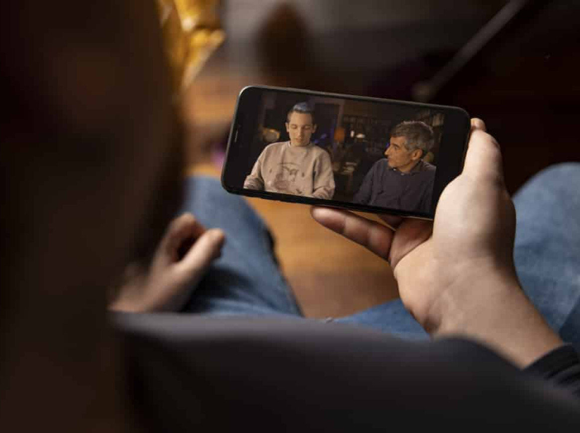 A person holds a smartphone while watching a video. The video shows two people sitting side by side, engaged in a conversation. The person on the left wears a light-colored sweater, and the person on the right wears a dark-colored shirt.