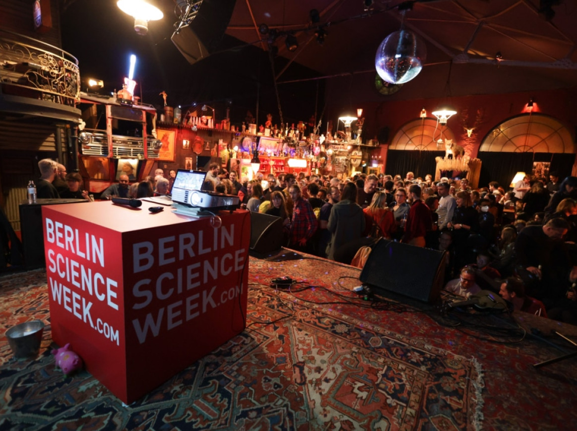 A crowded indoor venue with a diverse audience and a large red cube displaying the text "BERLIN SCIENCE WEEK.com." The venue has eclectic decor, including various lights, shelves with decorative items, and an illuminated disco ball hanging from the ceiling.