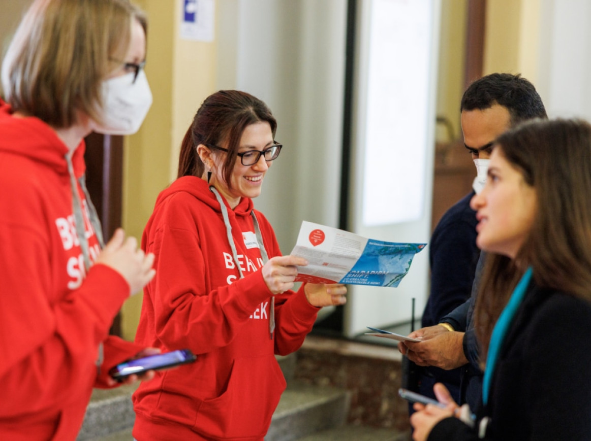 Two people wearing red hoodies and name tags engage in conversation with two others, one holding a smartphone, the other a pamphlet. One person in a red hoodie is masked and holding a smartphone, while the unmasked one is smiling warmly.